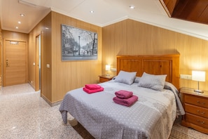 Hotelkamer met een houten lambrisering, een groot bed met een grijze sprei, twee nachtkastjes met lampen en een lijst met een stadsgezicht aan de muur.