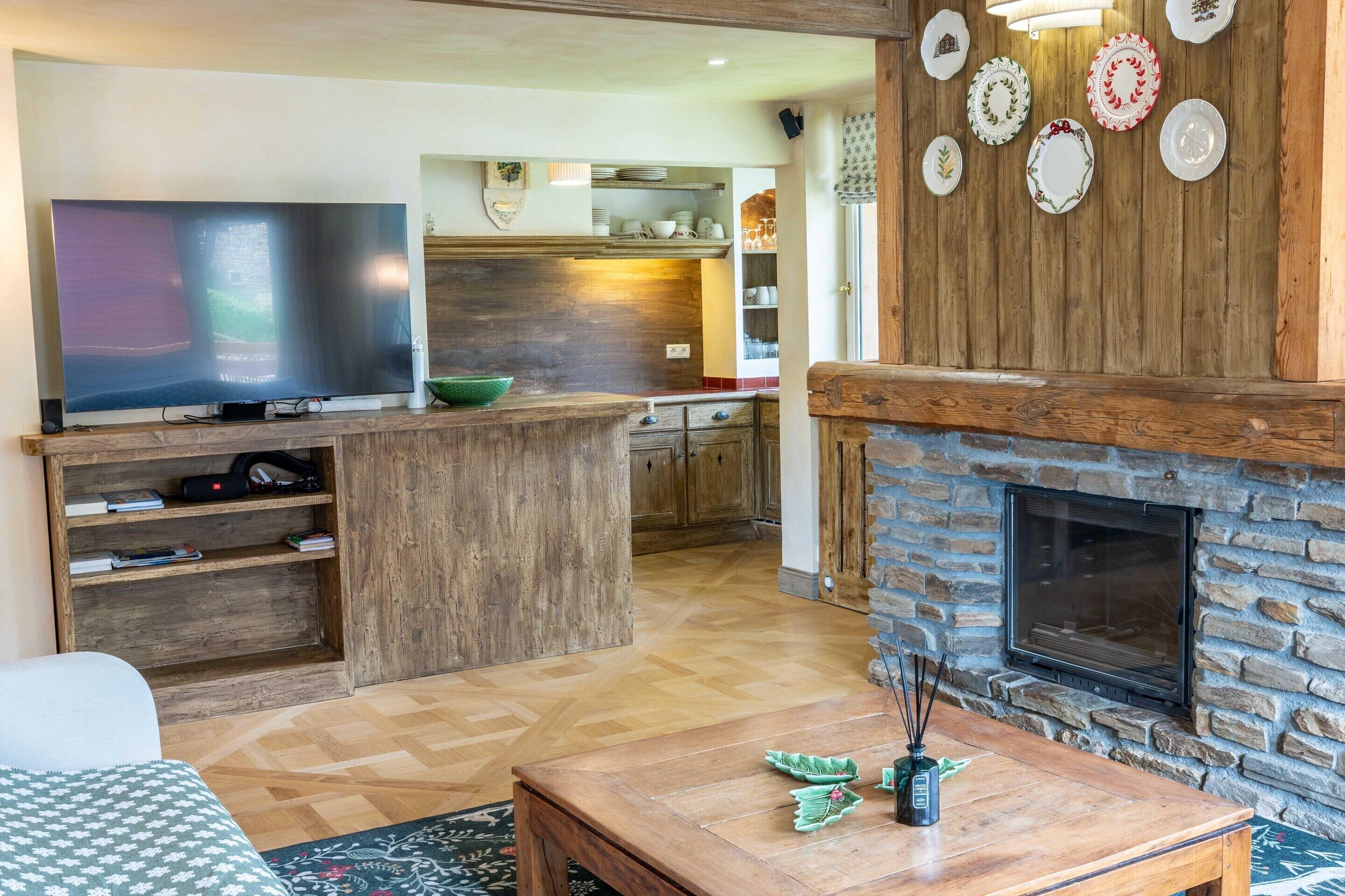 A living room with a TV, wooden entertainment unit, a fireplace, and a kitchen in the background.