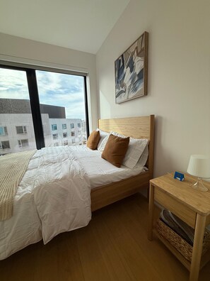 Ein Hotelzimmer mit einem großen Bett, einem Holz-Kopfteil, einem Nachttisch mit Lampe und Blick auf Gebäude durch ein Fenster.