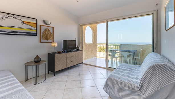 A modern living room with a sofa, a TV on a cabinet, a small table with a lamp, and a balcony with a view. - Studio 26m² “Marina de Sant Ambroggio” with Sea View, Terrace, Wi-Fi & Air Conditioning (Lumio)