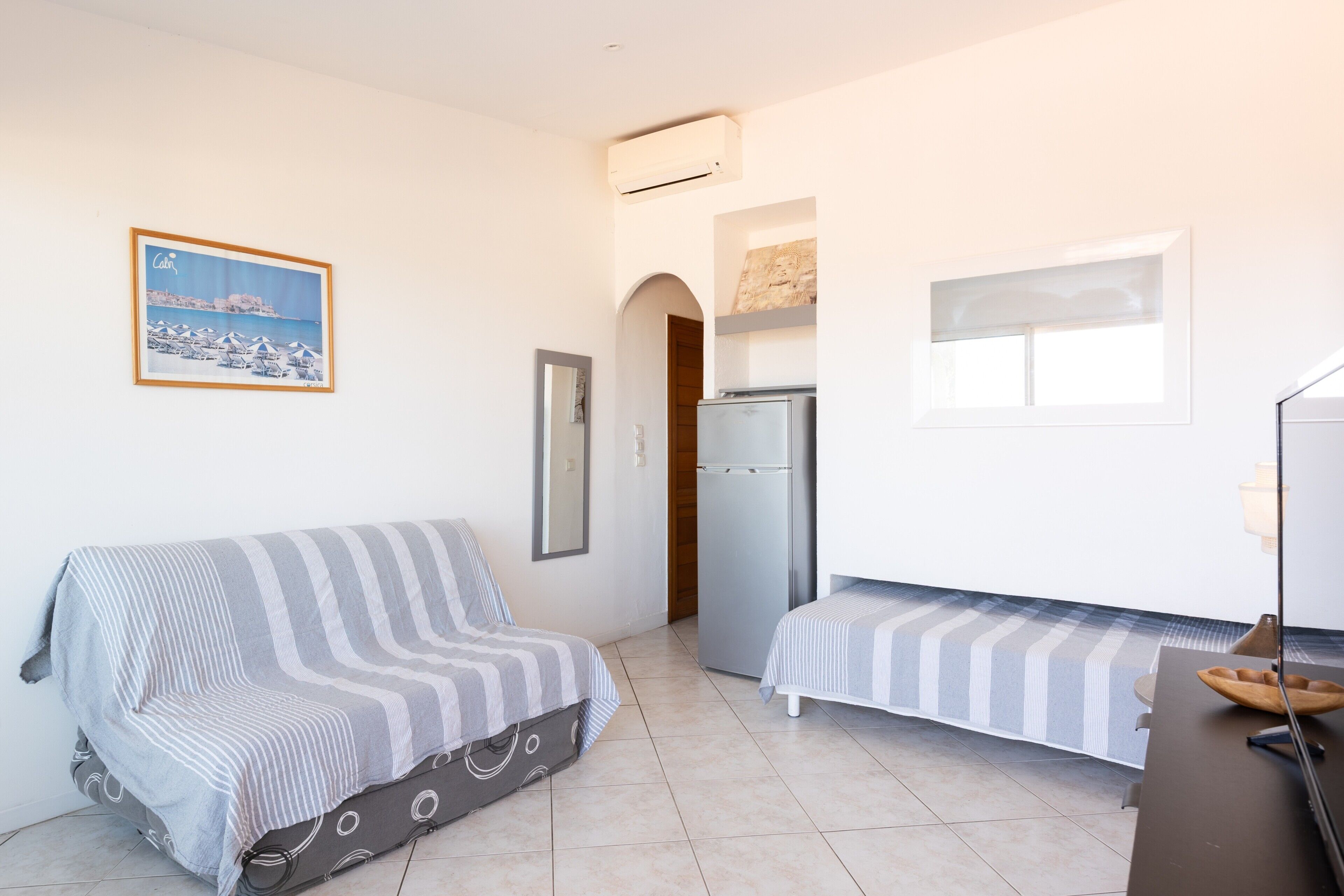 A compact room with a sofa, a small bed, a refrigerator, a window, and a picture on the wall.