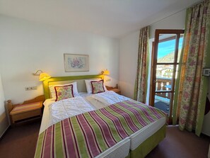 A double bed with striped bedding, two bedside lamps, a framed picture on the wall, and a window with floral curtains. - Fackler Apartments 7 Baumgarten – Bavarian Comfort & Mountain View in Tegernsee (Tegernsee)