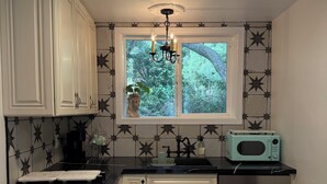 Una cocina con armarios blancos, encimera negra, ventana con vistas a vegetación, candelabro y microondas.