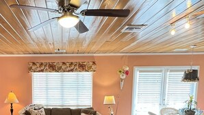 A living room with a wooden ceiling, a ceiling fan, a sofa, a coffee table, a dining area with a bar, and a kitchen counter. - Charming one floor, 2-bedroom house steps to the beach, private heated pool (Holmes Beach)