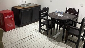 Eine kompakte Küche mit Spüle, einem Holzkasten und einem Esstisch mit vier Stühlen. Auf dem Boden ist ein roter Coca-Cola Kühlschrank zu sehen.