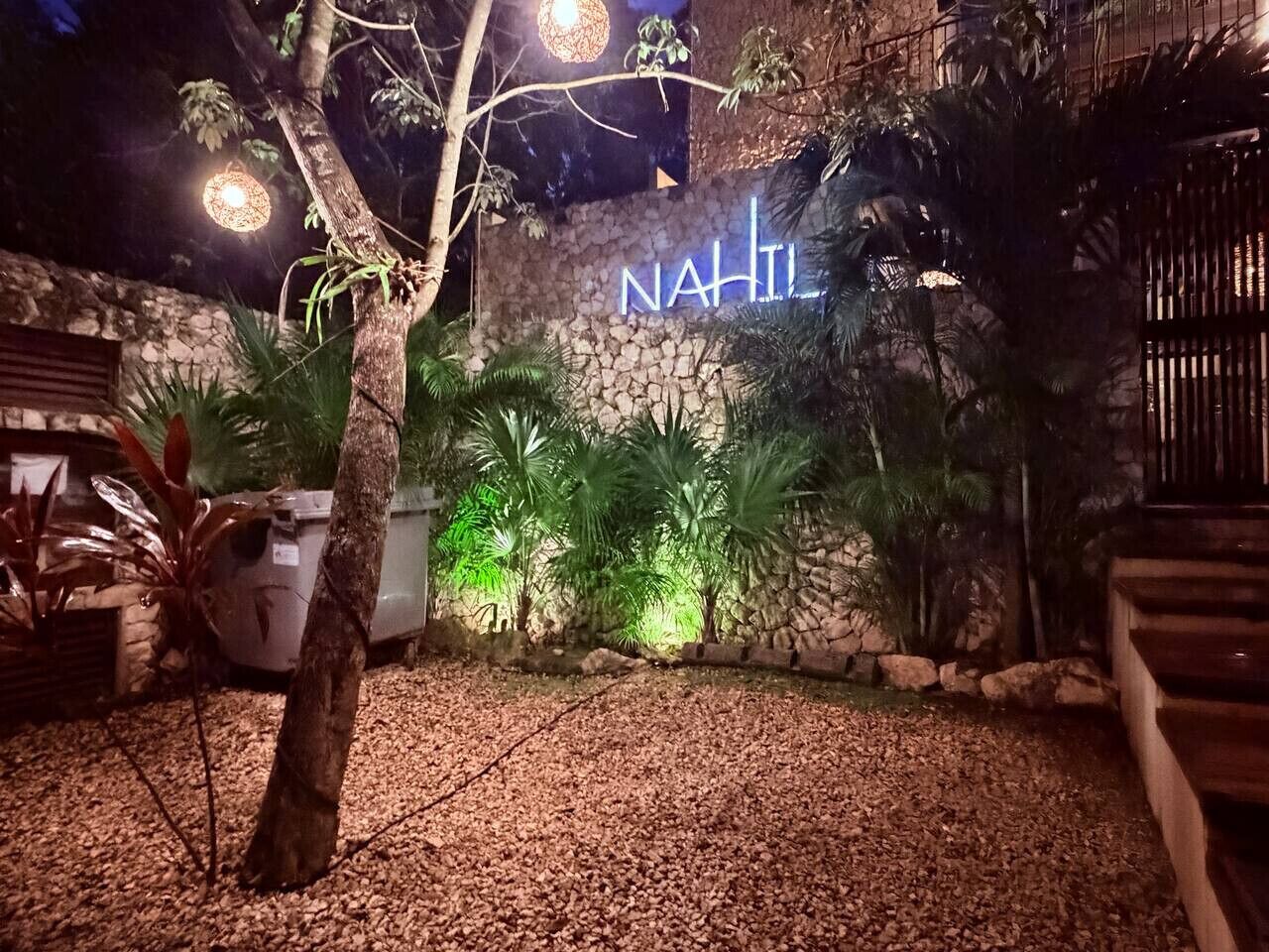 Kivimuuri, jossa on neonvalomainos, jossa lukee ”NAHIL”, ja puu etualalla.