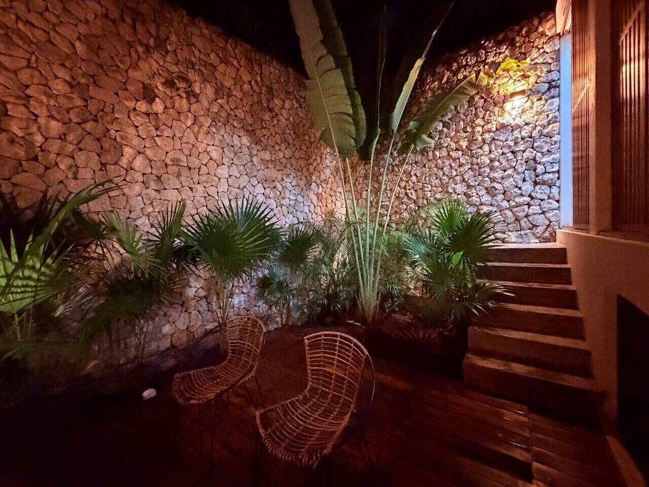Een stenen muur met een trap die omhoog leidt, omringd door plantenpotten en rieten stoelen.