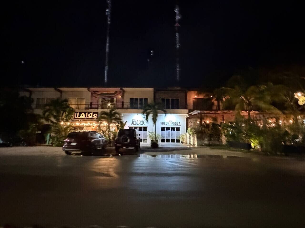 A building with illuminated signage, a parking area, and a reflection on the wet ground.