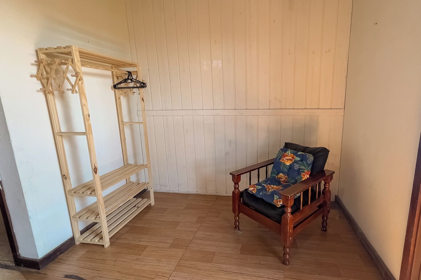 Une pièce avec un plancher en bois, une chaise en bois dotée d’un coussin à motifs floraux et une étagère en bois pourvue de crochets.