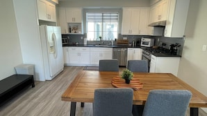 A modern kitchen with white cabinets, a wooden dining table with four chairs, and a window with blinds. - Modern New 3BR/2.5BA Home in SGV • Stylish, Spacious & Fully Furnished (South El Monte)