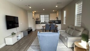 A modern living room with a grey sofa, a TV, and a dining area with a table and chairs. - Modern New 3BR/2.5BA Home in SGV • Stylish, Spacious & Fully Furnished (South El Monte)