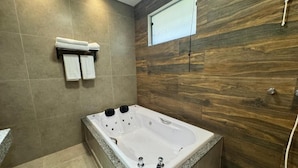 A modern bathroom with a large bathtub, wooden wall paneling, and a window with blinds. - Pousada Karandá (Serra Negra)