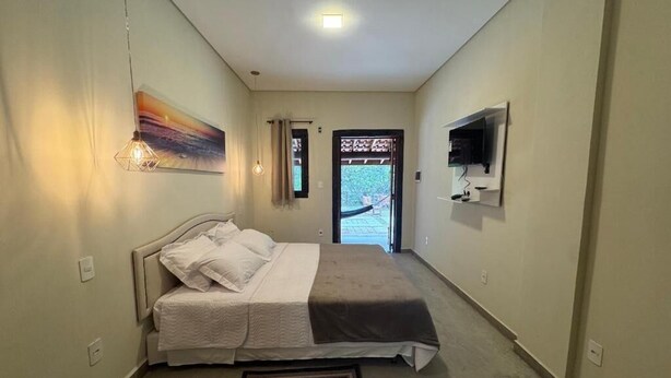 A bedroom with a bed, a painting, a TV, and a view through the door. - Pousada Karandá (Serra Negra)