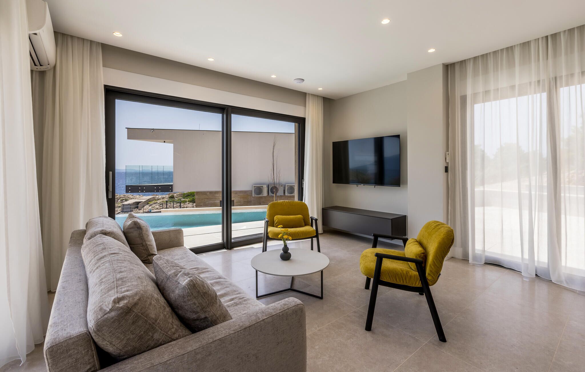 A modern living room with a sofa, two yellow chairs, a small round table, and a TV. A large window offers a view of a pool and the sea.