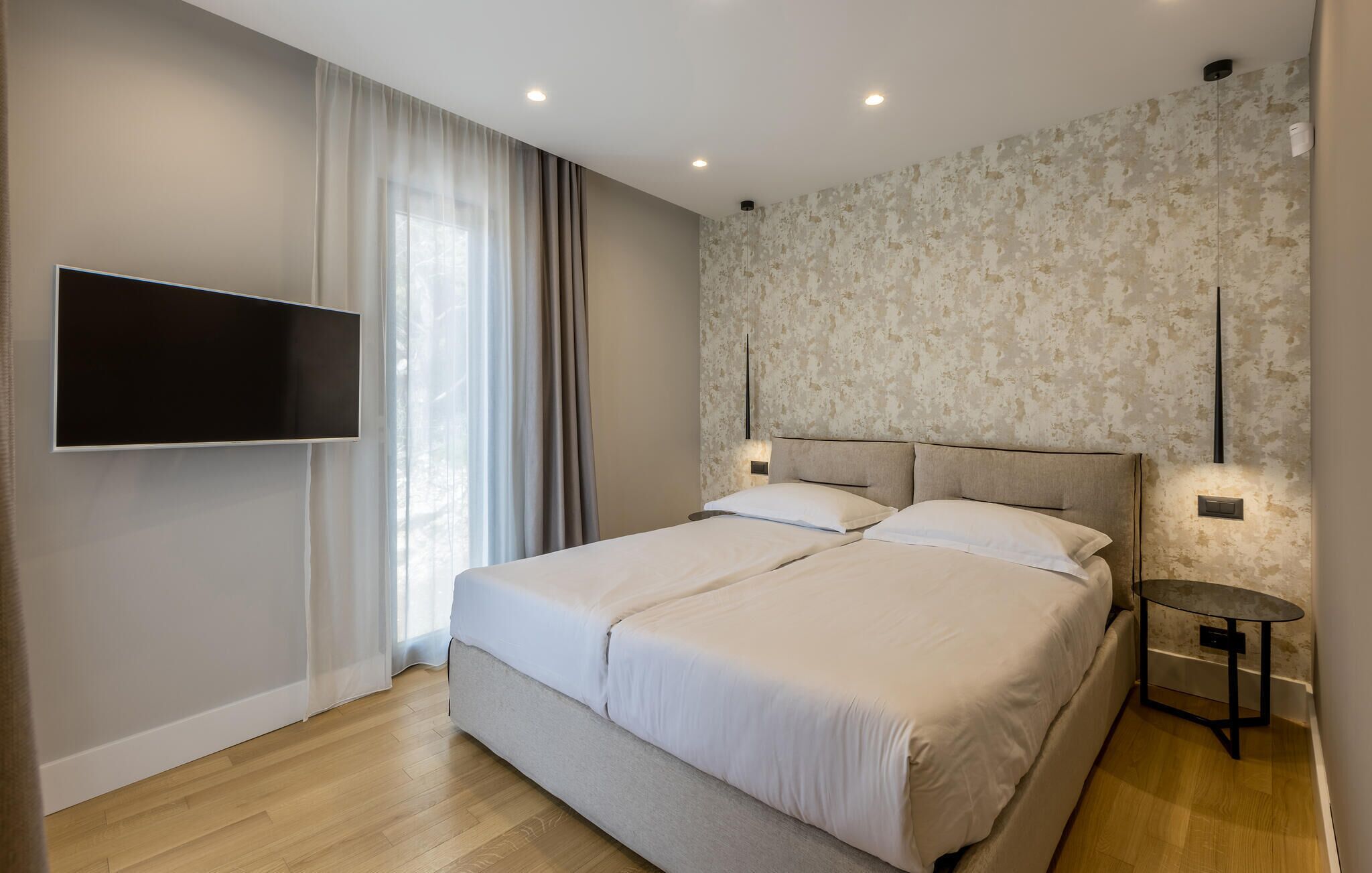 A modern bedroom with a double bed, a TV mounted on the wall, and a small table with a lamp.