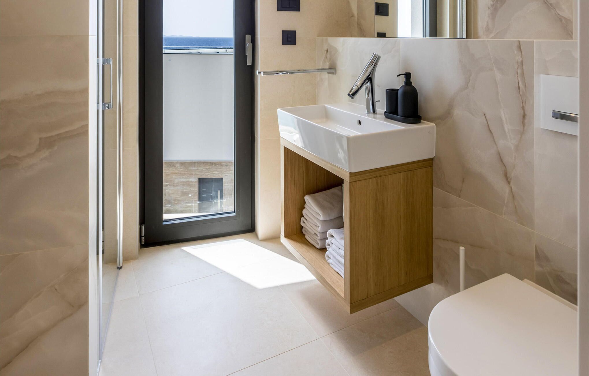 A modern bathroom with a white sink, wooden cabinet, and a view of the sea through a glass door.