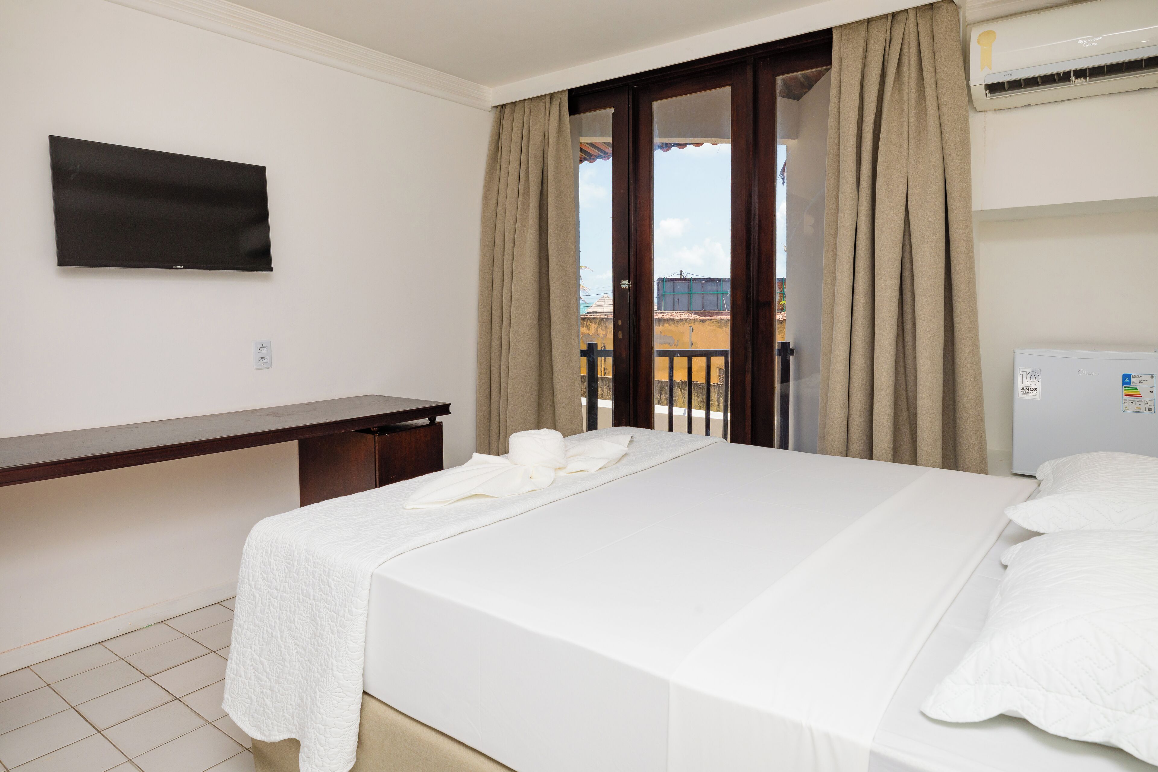 A hotel room with a bed, a desk, a TV mounted on the wall, and an air conditioning unit.