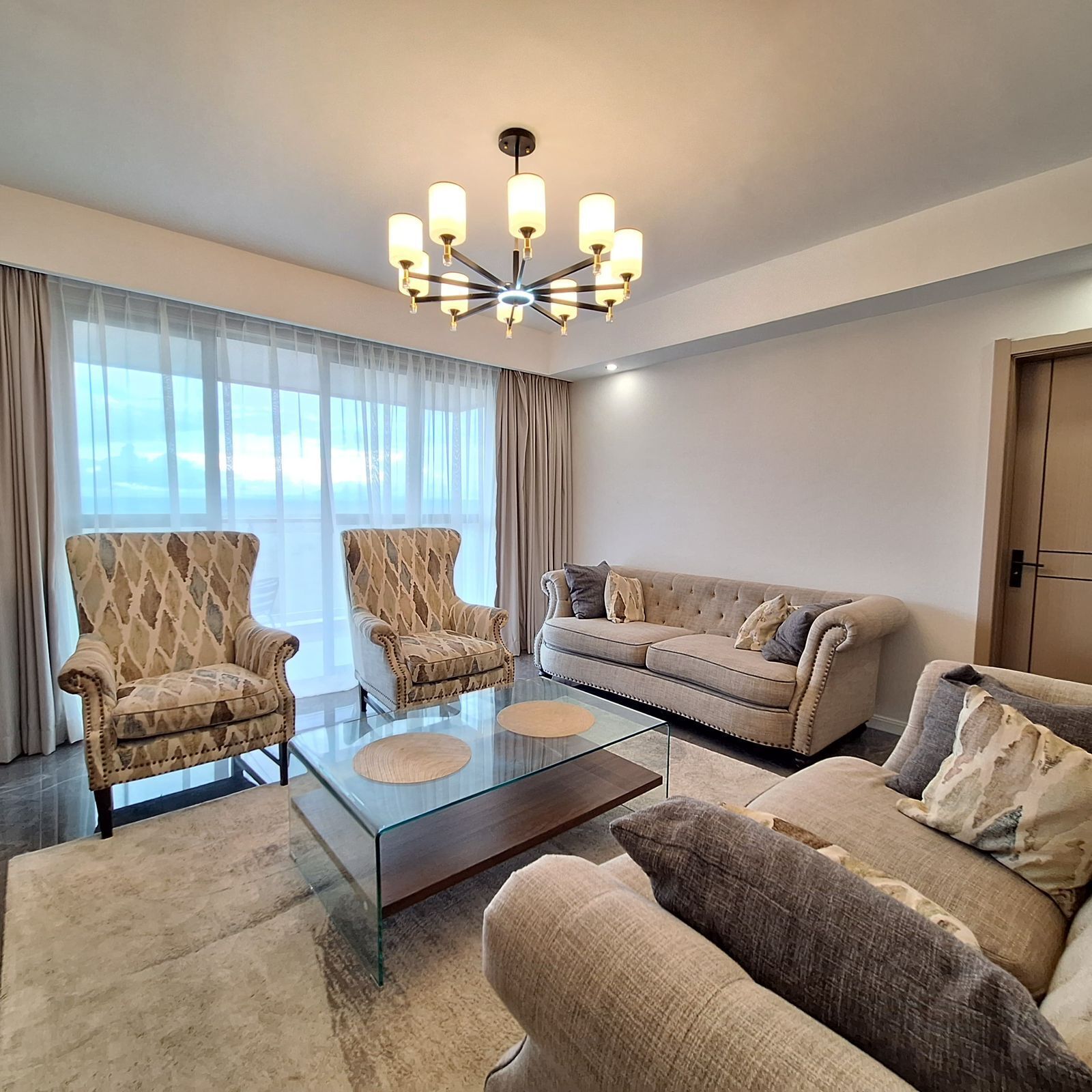 A modern living room with a glass coffee table, two armchairs, a sofa, and a chandelier.