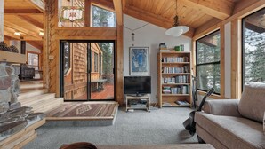 A cozy cabin interior with a stone fireplace, wooden shelves, a bookshelf, and a large window offering a view of the outdoors. - Cozy Donner Lake Cabin w/ Kayaks, Forest Views, Fireplace & Quick Ski Resort Access Donner Lake Refuge by Avantstay (Truckee)