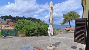 A coastal view with a beach umbrella, a stone wall, and a building with a brown shutter. - Antunovic / Antunovic 1 (Veli Lošinj)