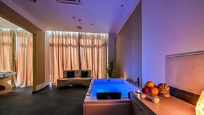 Et moderne spa-rum med et stort boblebad, et siddeområde med sofa og stole, samt en bordplads med frugt og en lampe.