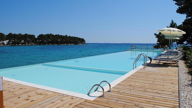 A clear blue pool with wooden deck, lounge chairs, and umbrellas, overlooking a calm sea and a tree-lined shore. - Cozy apartment in Biograd with sauna (Biograd)