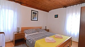Een slaapkamer met een houten plafond, een bed met beddengoed met een patroon, twee nachtkastjes met lampen, en een raam met witte gordijnen.