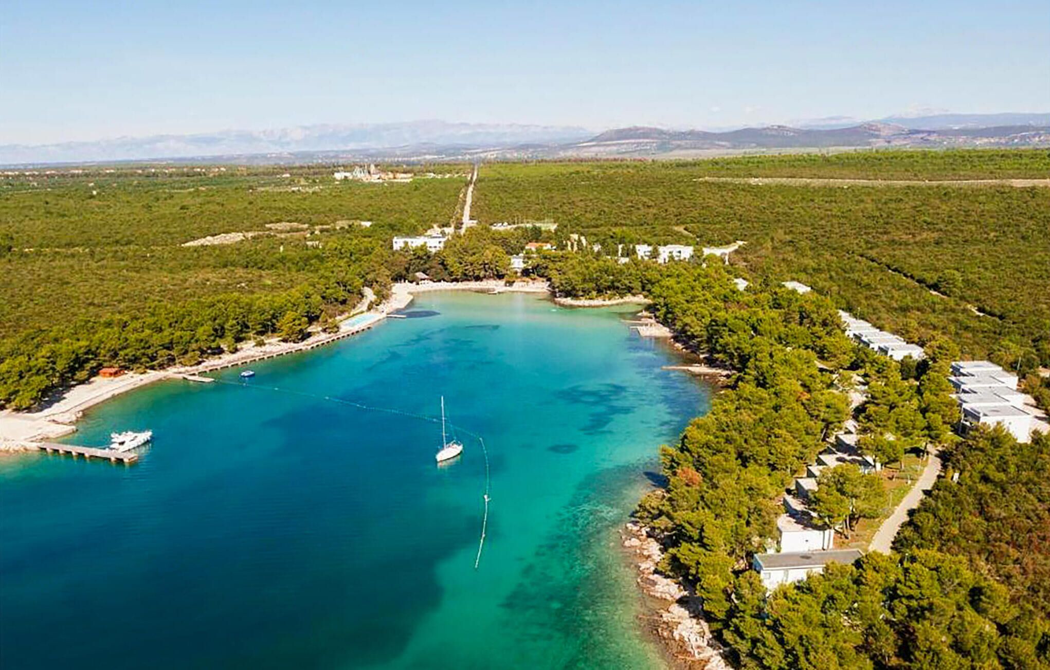 Une vue aérienne d’une zone côtière avec une baie d’un bleu limpide, entourée d’une végétation luxuriante et d’un petit village avec des bâtiments le long de la côte.