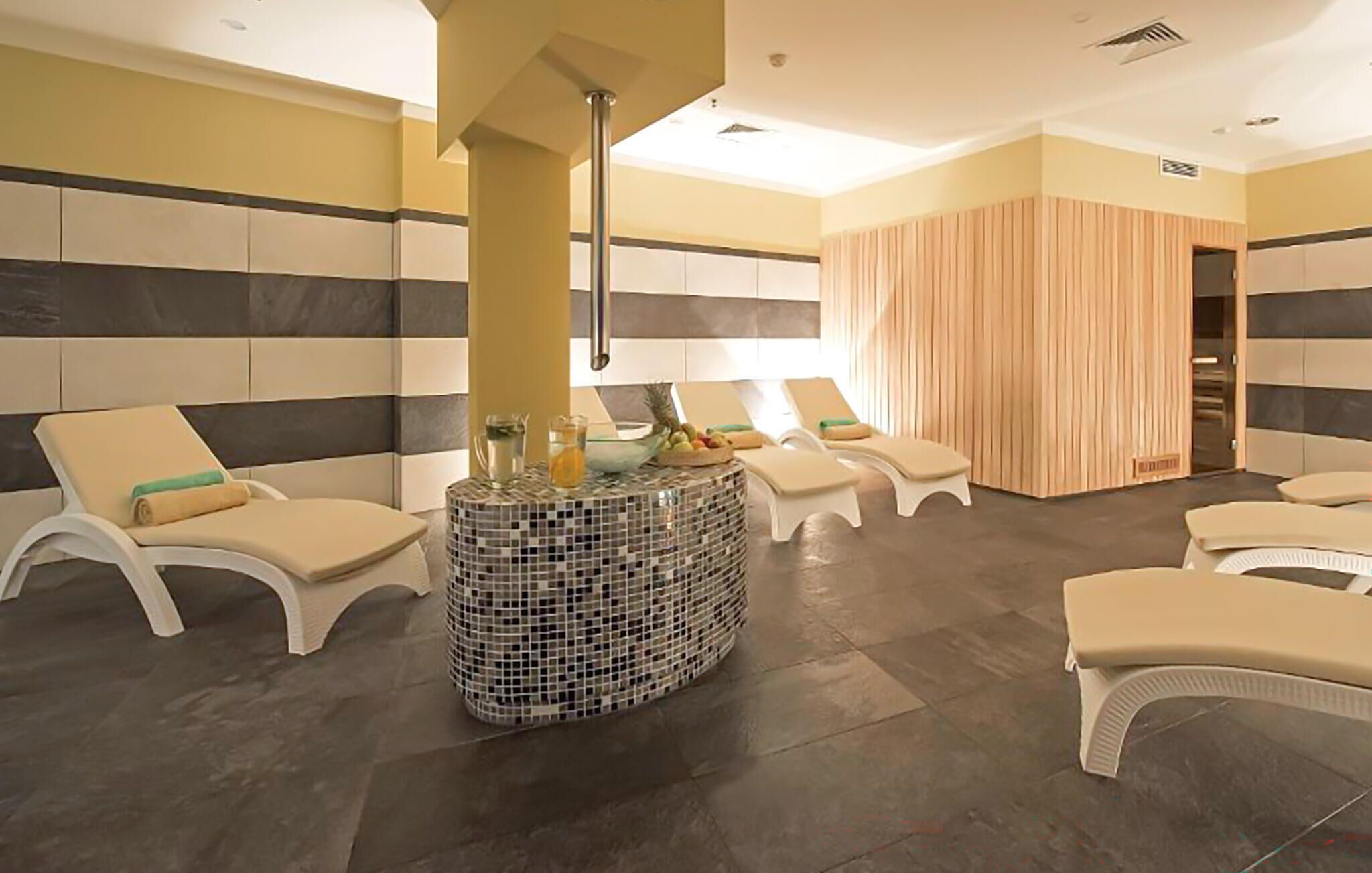 Un área de spa con varias sillas blancas acolchadas, una mesa de mosaicos con una bebida y fruta, y un panel de pared de madera.