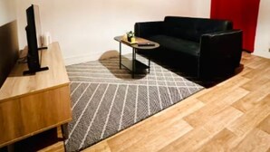 A modern living room with a wooden floor, a black sofa, a small coffee table, and a TV on a wooden stand. There are three framed pictures on the wall and a red curtain in the background.