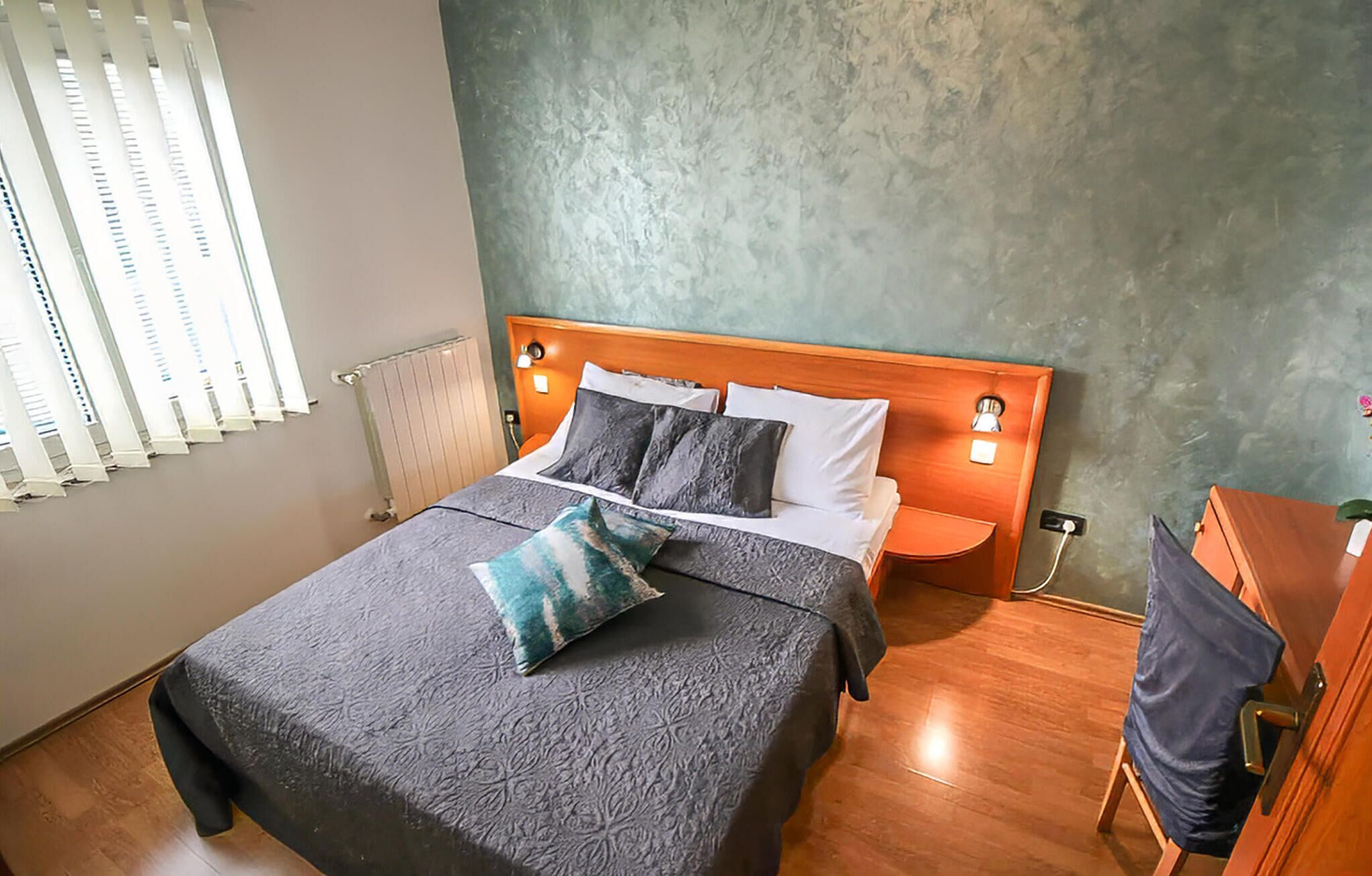 A neatly made bed with a grey bedspread and pillows, a wooden headboard, bedside tables with lamps, and a radiator on the wall.