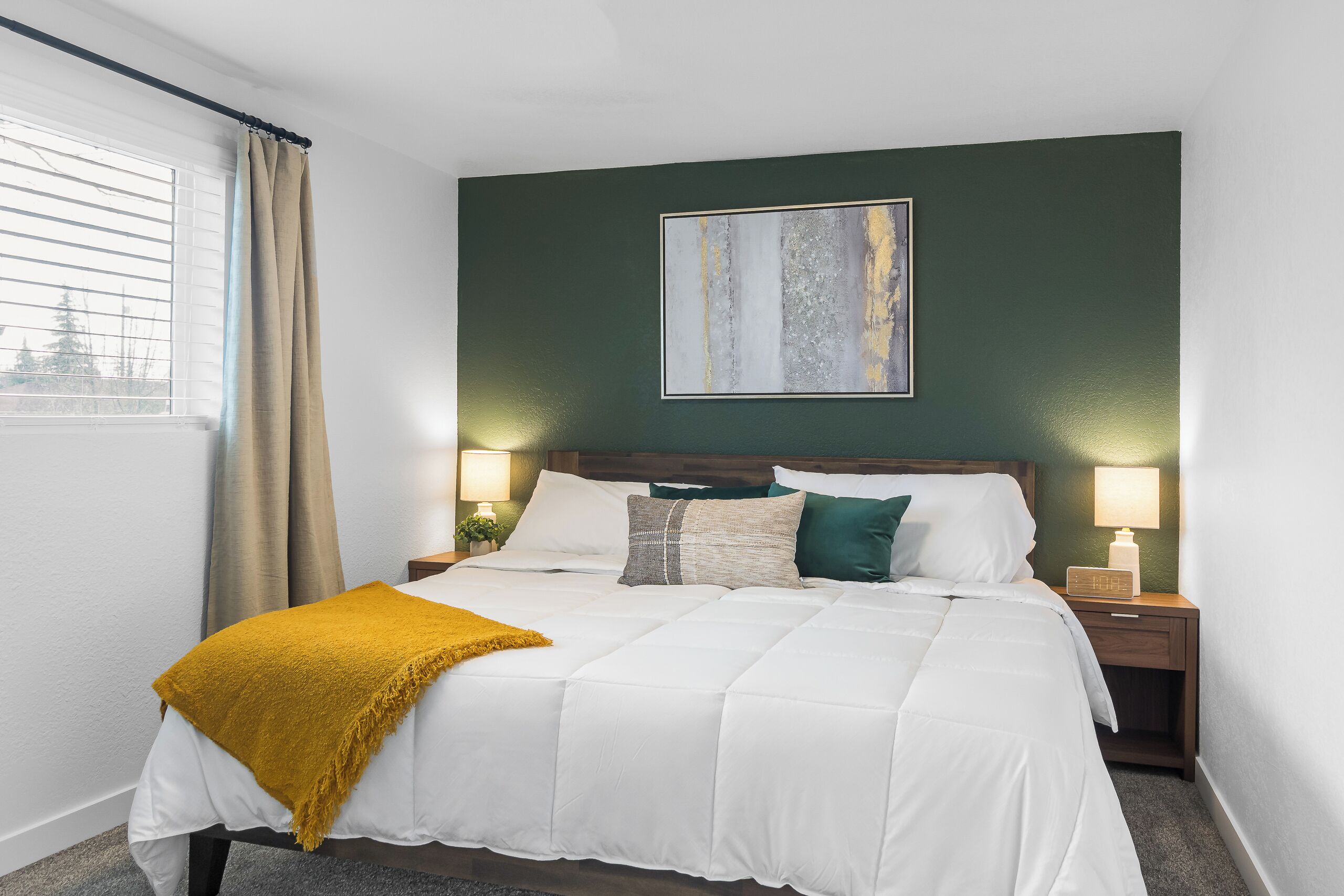 A neatly made bed with a mustard throw blanket, a headboard with a painting, two bedside lamps, and a window with curtains.