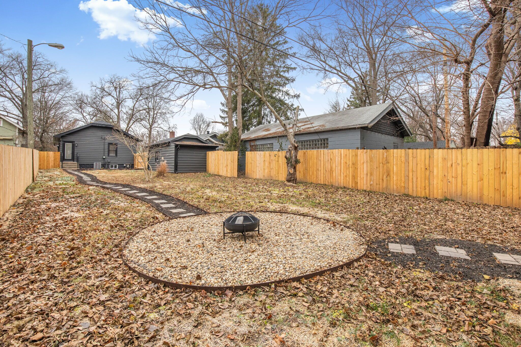 A backyard with a circular gravel area, wooden fence, and a metal fire pit.