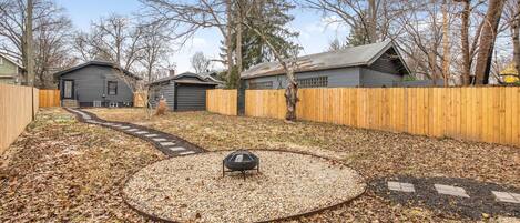 A backyard with a circular gravel area, wooden fence, and a metal fire pit.