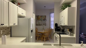 A modern kitchen with white cabinetry, a dining area with wooden chairs, a refrigerator, a coffee maker, and a sink. - Legends Country Club Social 2/2 Condo Overlooking Hole #1, Close to Country Club (Fort Myers)