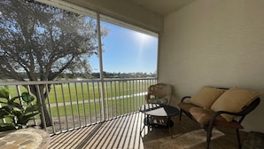 A balcony with a view of a grassy field, a wicker chair, a cushioned sofa, and a small table. - Legends Country Club Social 2/2 Condo Overlooking Hole #1, Close to Country Club (Fort Myers)