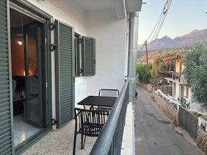 A balcony with a table and chairs, a view of a mountain range, and a street with a fence and a building with a balcony. - Anemeli 2 bedroom Apartment, Stoupa, 50m to beach. Stunning Sea & Mountain views (Stoupa)