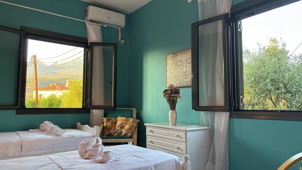 A hotel room with two beds, a window with a view of greenery and a building, a bedside table with a vase of flowers, and an air conditioning unit. - Anemeli 2 bedroom Apartment, Stoupa, 50m to beach. Stunning Sea & Mountain views (Stoupa)