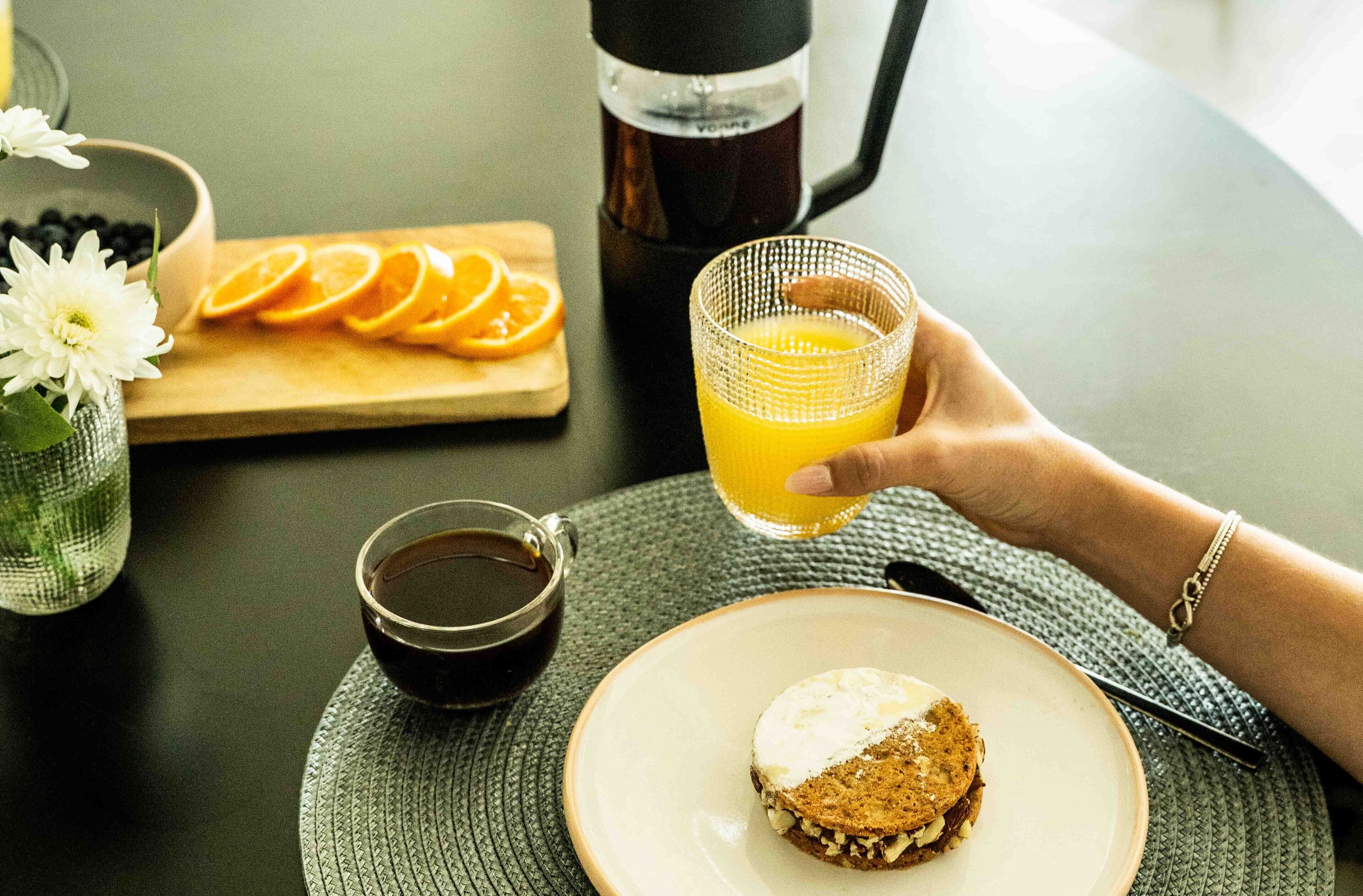 Desayuno con taza de café, rodajas de naranja, vaso de zumo de naranja y un sándwich en un plato.