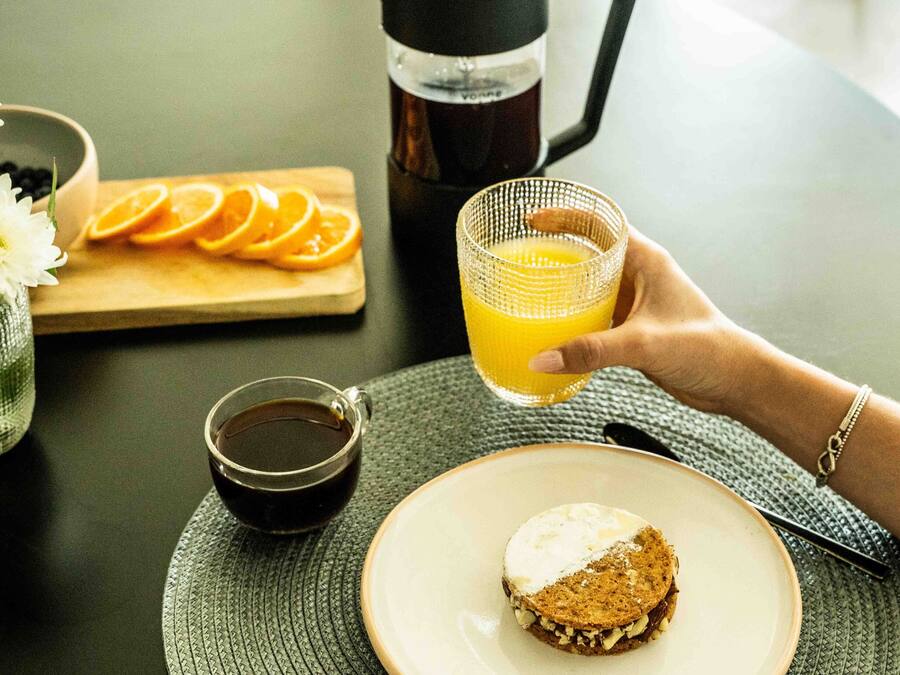 Desayuno con taza de café, rodajas de naranja, vaso de zumo de naranja y un sándwich en un plato.