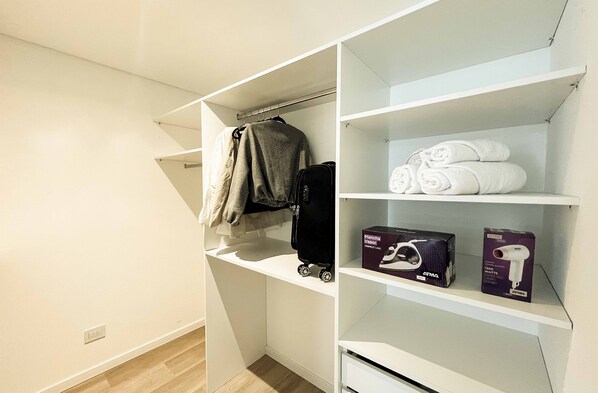 A white closet with shelves, a hanging rod, and a suitcase. - RentUp Suites Palmas Park Buenavista (Victoria)