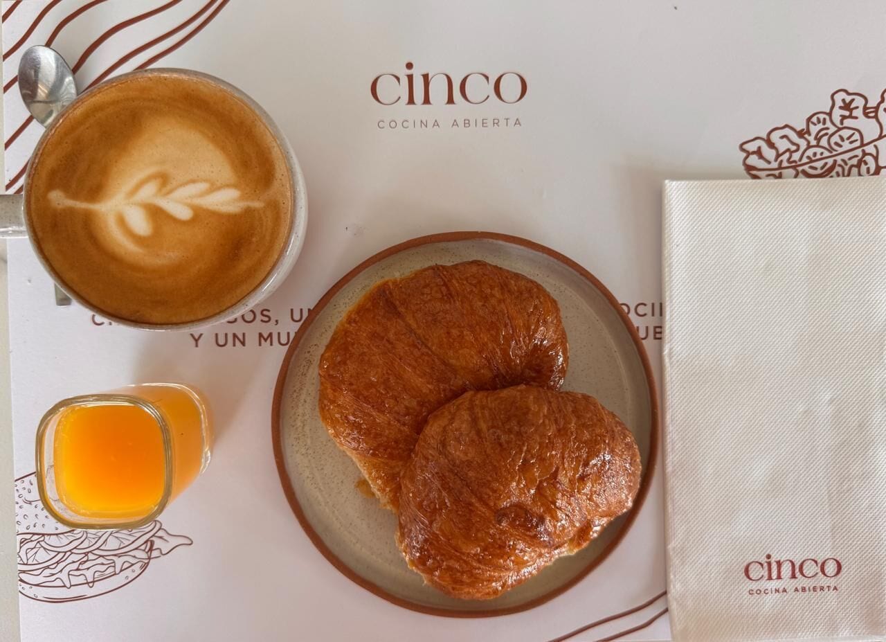 Un desayuno con croissants, café y un vaso de zumo de naranja sobre una mesa con servilleta y menú.