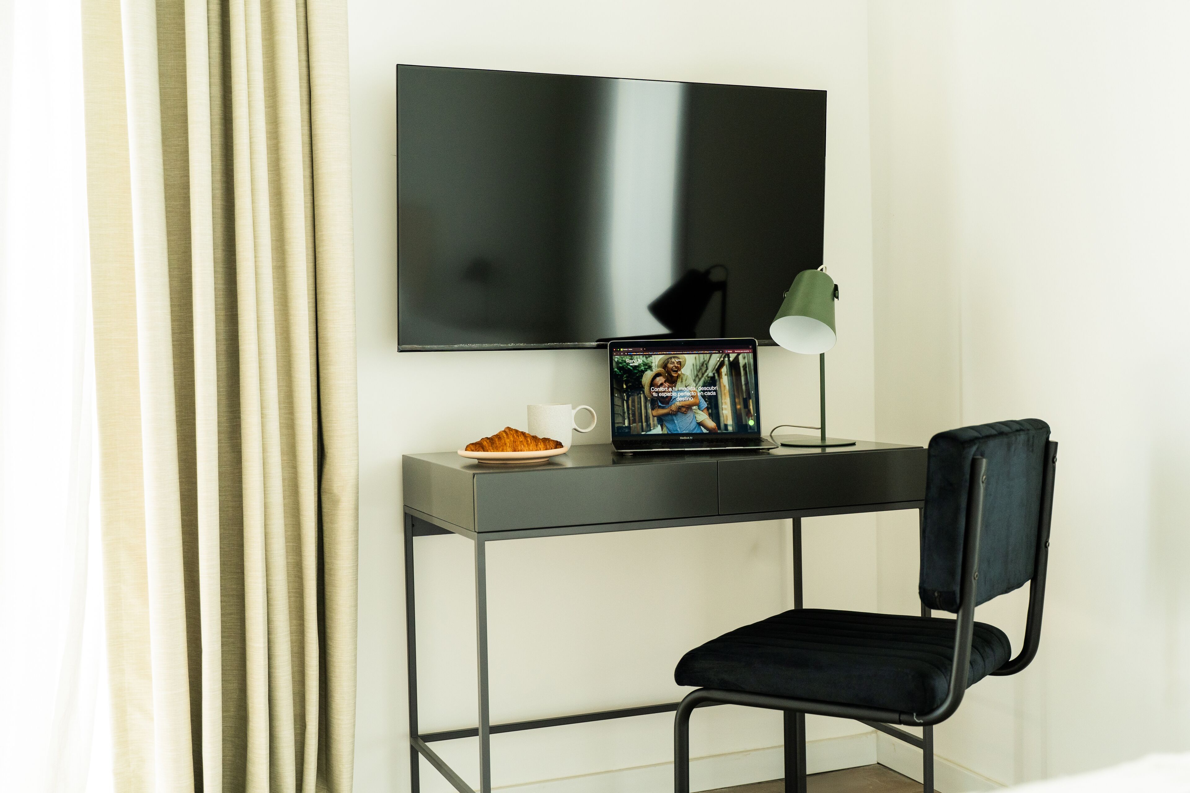 A modern room with a flat-screen TV mounted on the wall, a desk with a laptop, a lamp, and a chair.