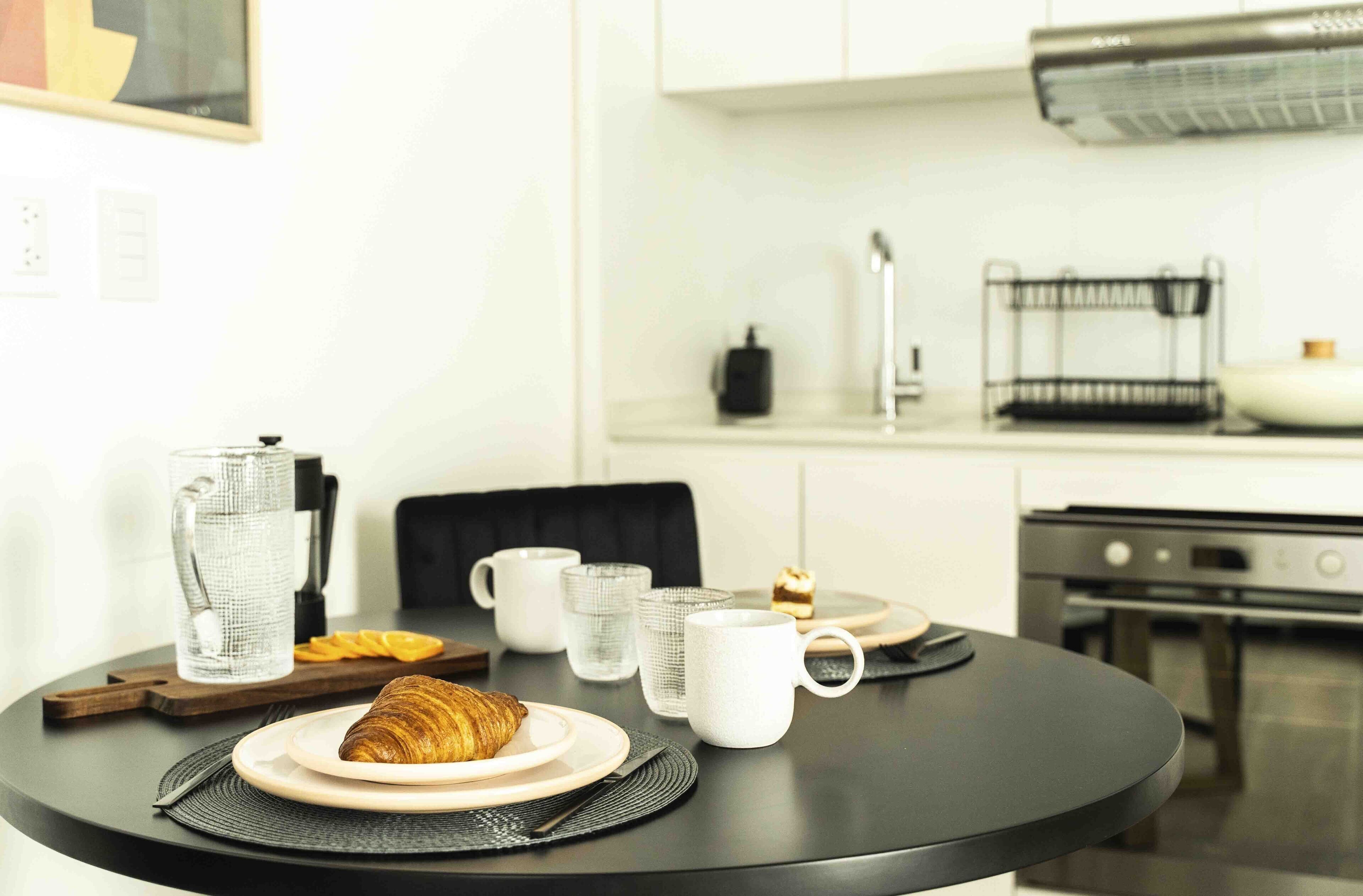 Un desayuno con un croissant, tazas de café y una tetera sobre una mesa redonda.