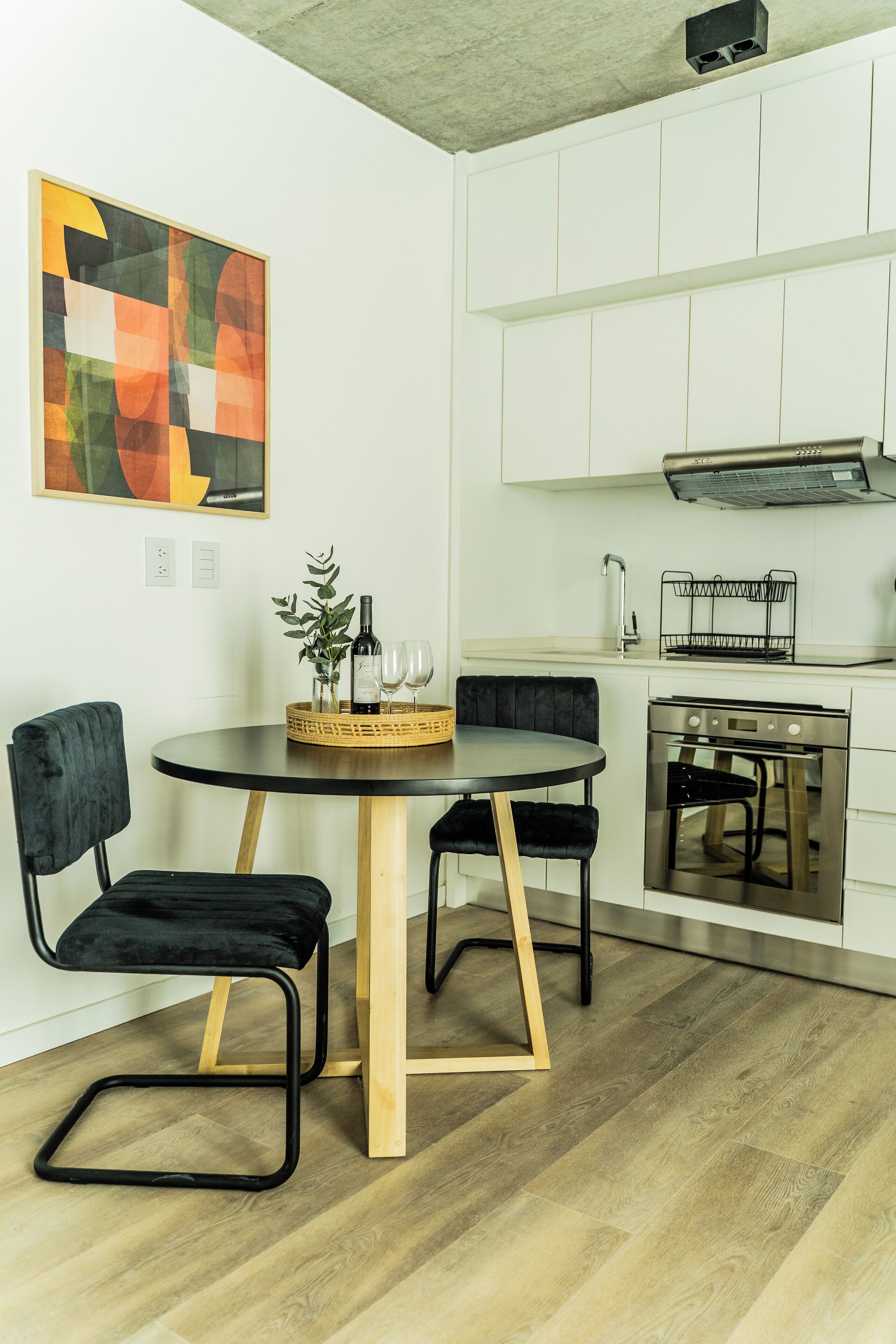 Una cocina moderna con gabinetes blancos, horno empotrado y comedor con una mesa redonda y sillas negras.