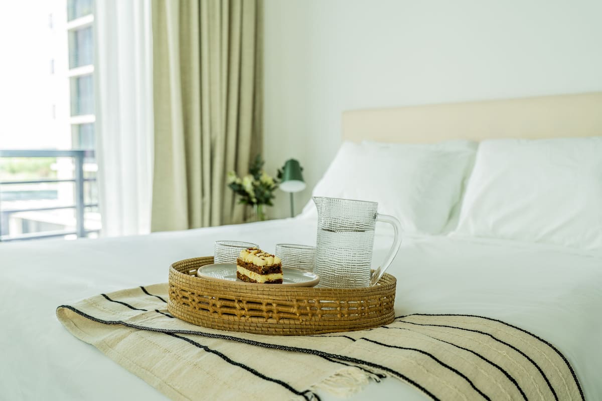A neatly made bed with a white comforter, a woven tray with a dessert and glasses, and a view of a balcony with curtains.