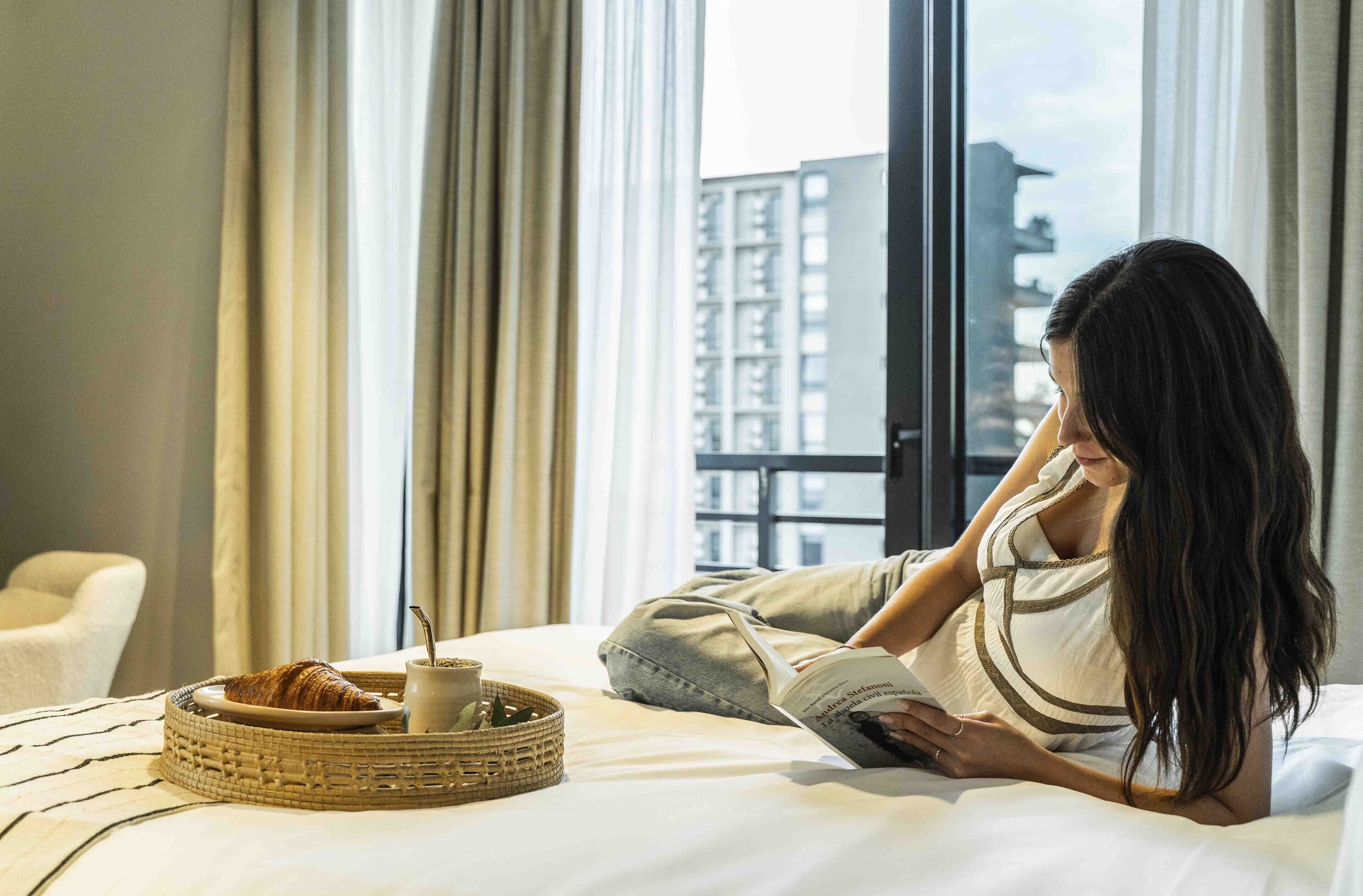 Eine Person liegt im Bett, neben einem Croissant und einer Tasse, und liest ein Buch. Durch das Fenster ist ein Blick auf die Stadt möglich.