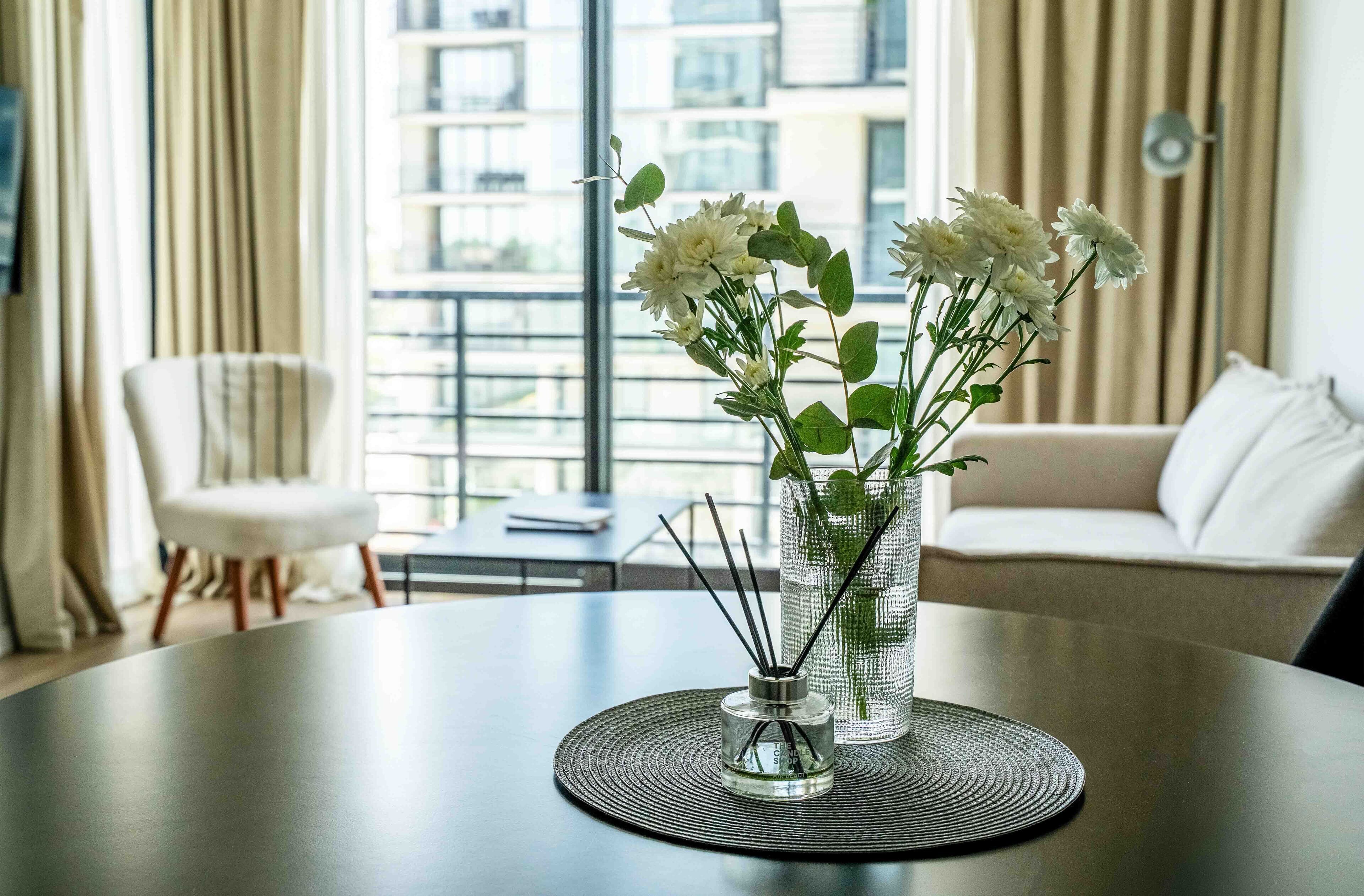 Una sala de estar moderna con una mesa redonda, un florero con flores y vistas a un paisaje urbano a través de amplios ventanales.