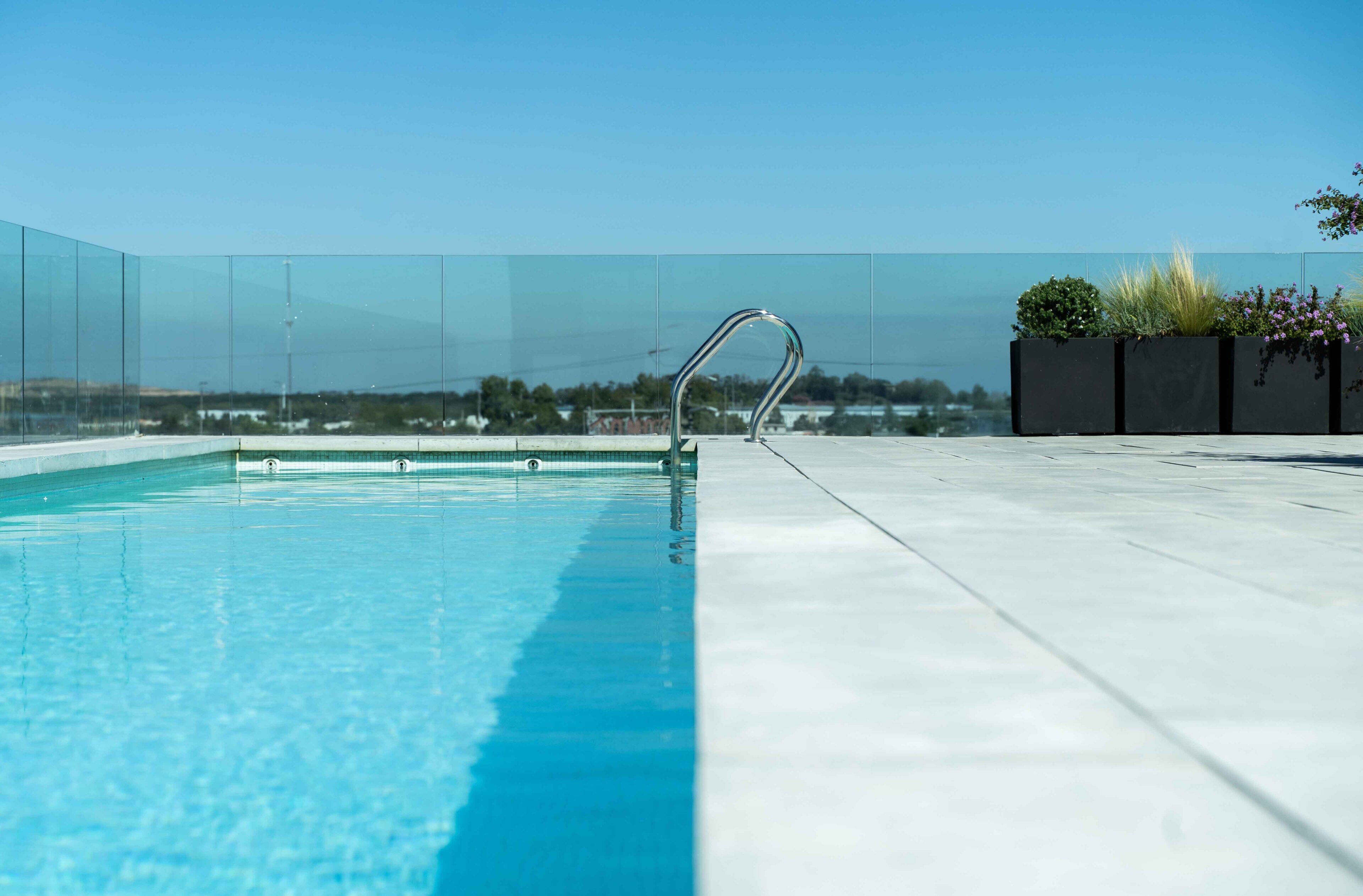 Una piscina azul cristalina con una escalera de metal, rodeada por una barandilla de vidrio y un macetero con plantas.