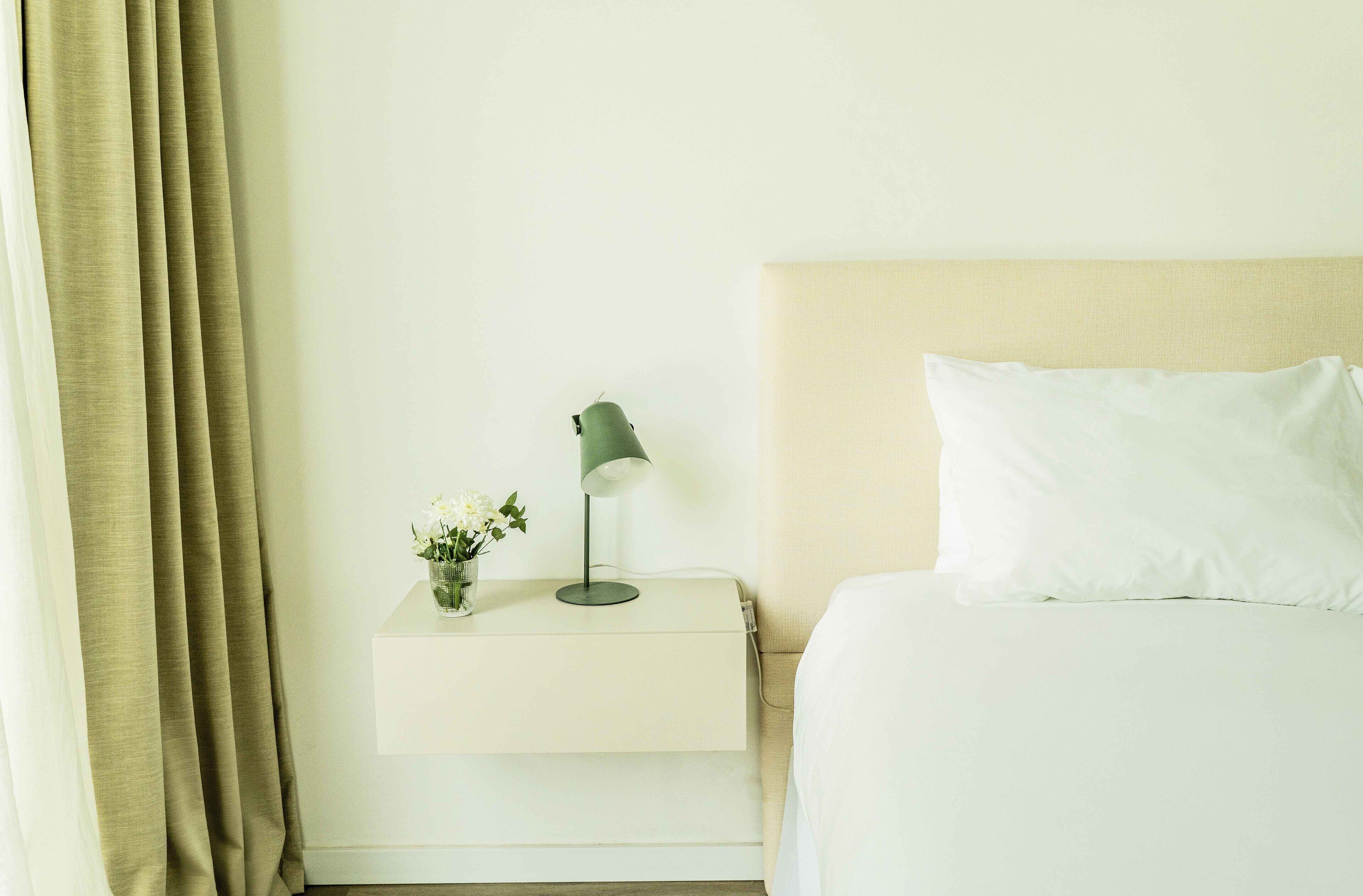 A neatly made bed with white linens, a bedside lamp, and a small vase with flowers on a white nightstand.